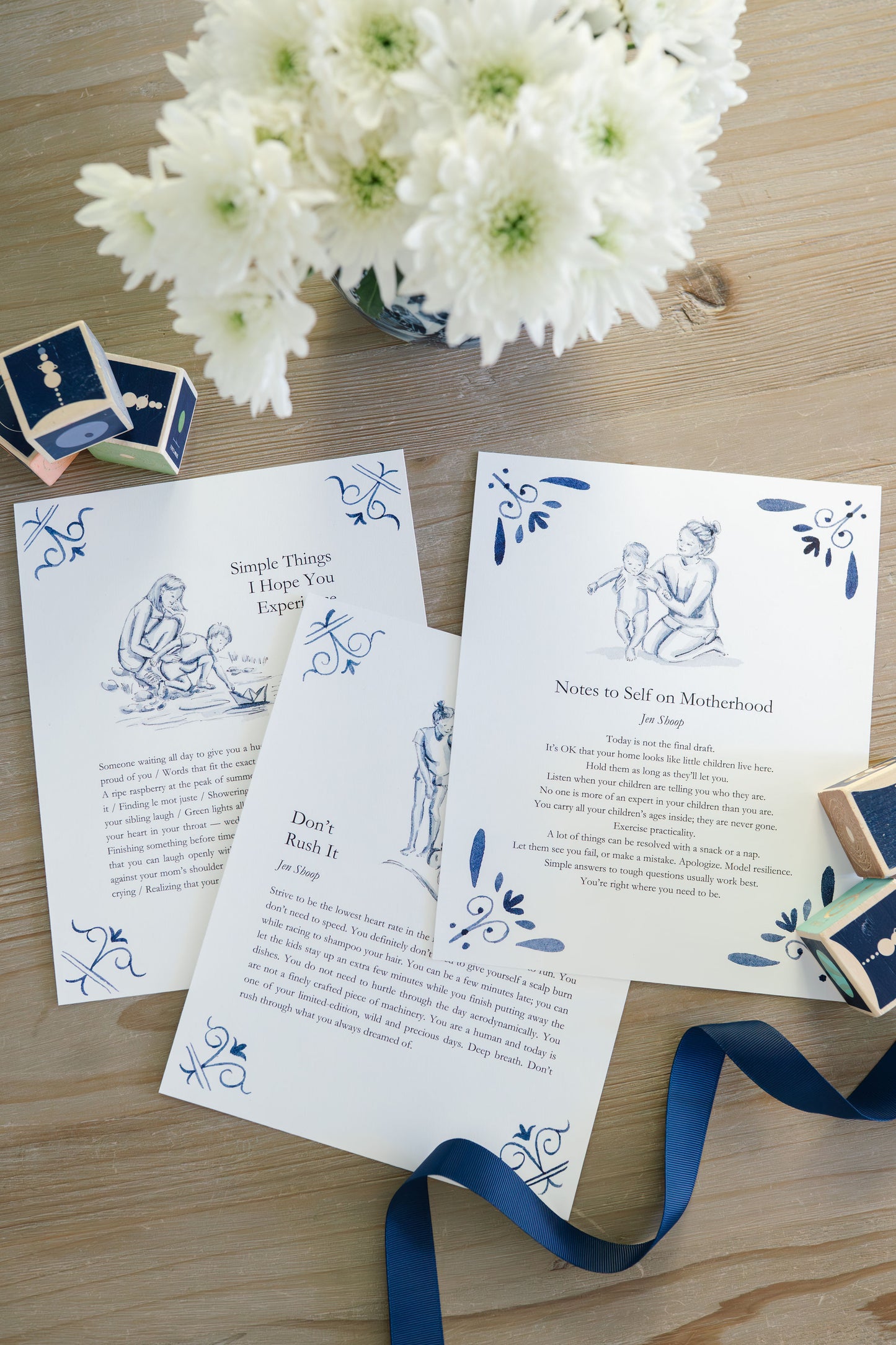 Three blue-and-white art prints—Simple Things, Don’t Rush It, and Notes to Self on Motherhood—styled on a wood table with flowers.